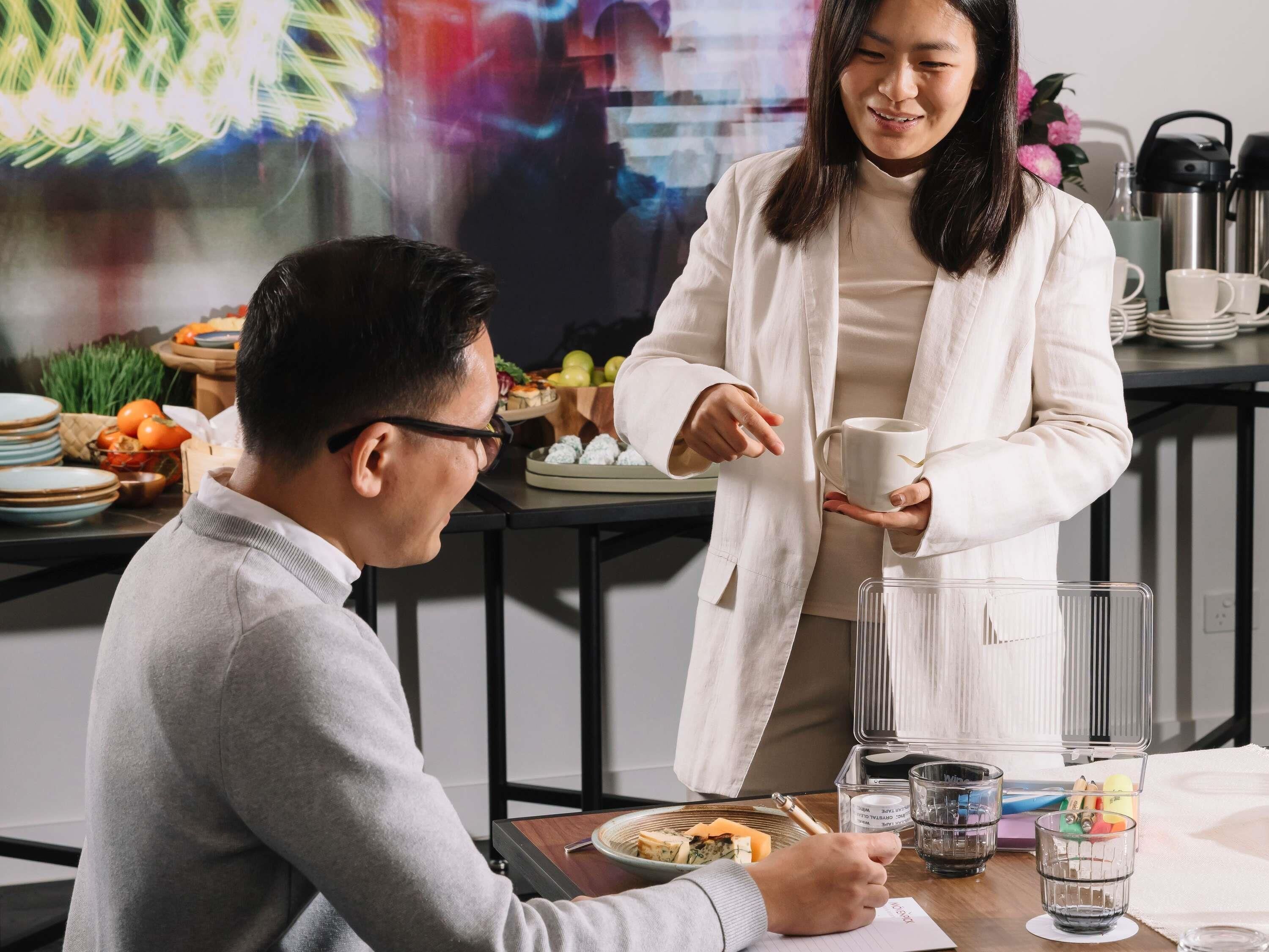 A person in a white coat holding a cup, seated at a table with plates, a thermos, and a glass.