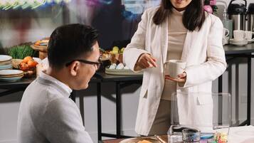 A person in a white coat holding a cup, seated at a table with plates, a thermos, and a glass.