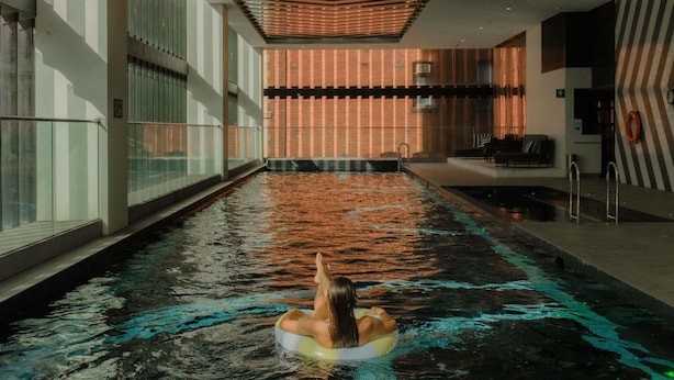Una piscina con una persona flotando en un flotador, rodeada de paredes de vidrio y un diseño de techo moderno.