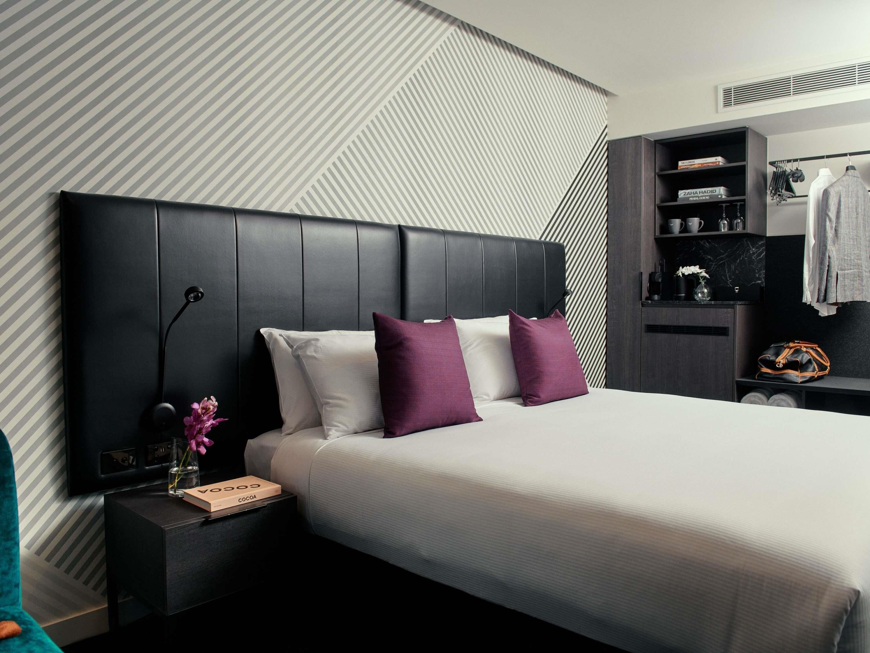Una habitación de hotel moderna con una cama grande, una mesita de noche, un armario y un estante empotrado con ropa y artículos de aseo.