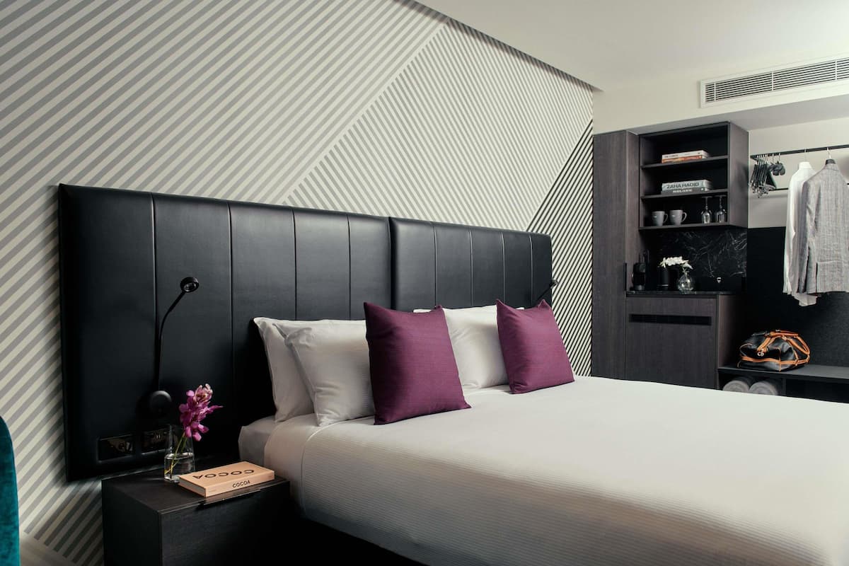 Una habitación de hotel moderna con una cama grande, una mesita de noche, un armario y un estante empotrado con ropa y artículos de aseo.
