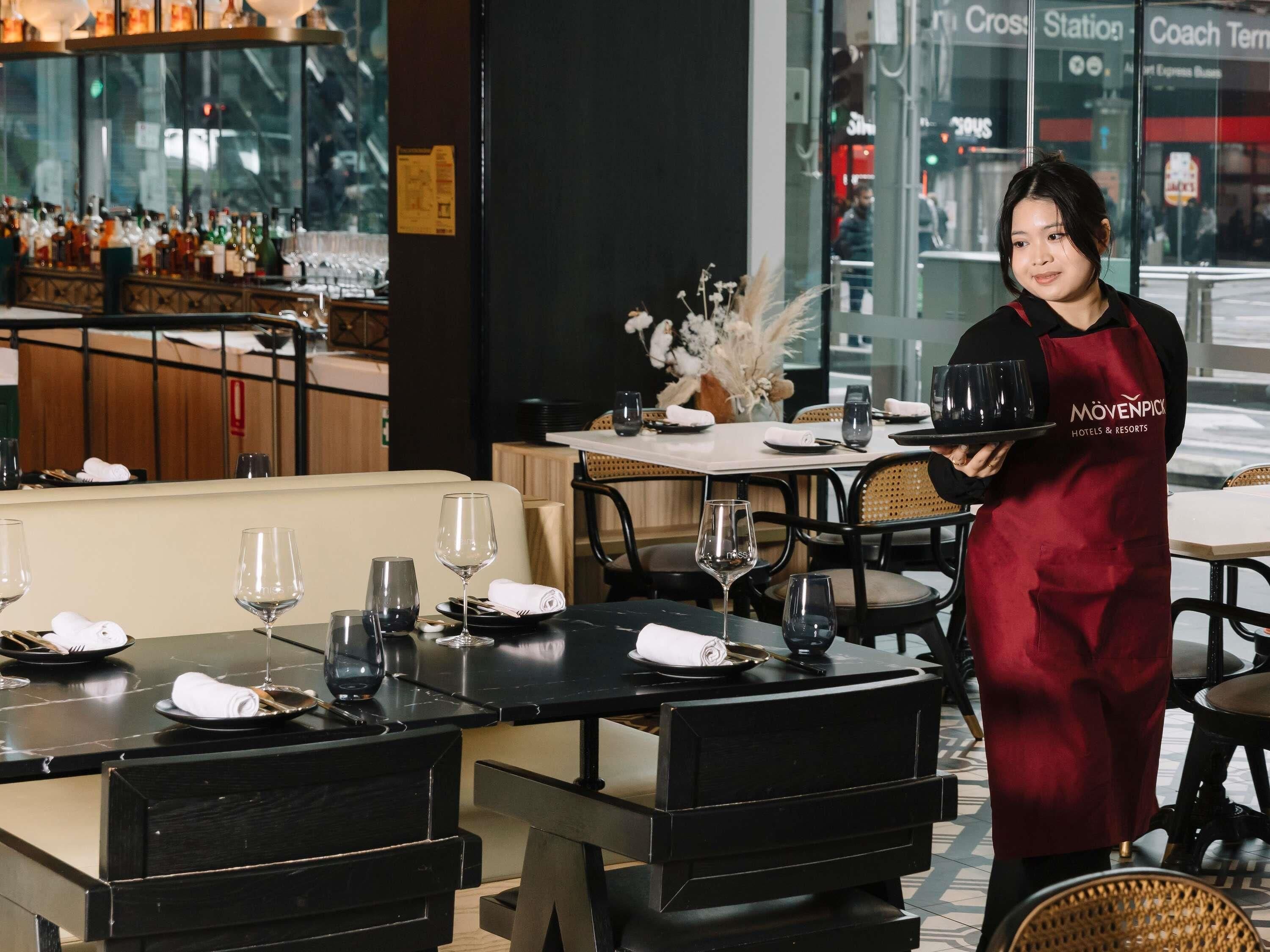 A restaurant setting with tables set for dining, a bar area with bottles, and a waiter carrying a tray.