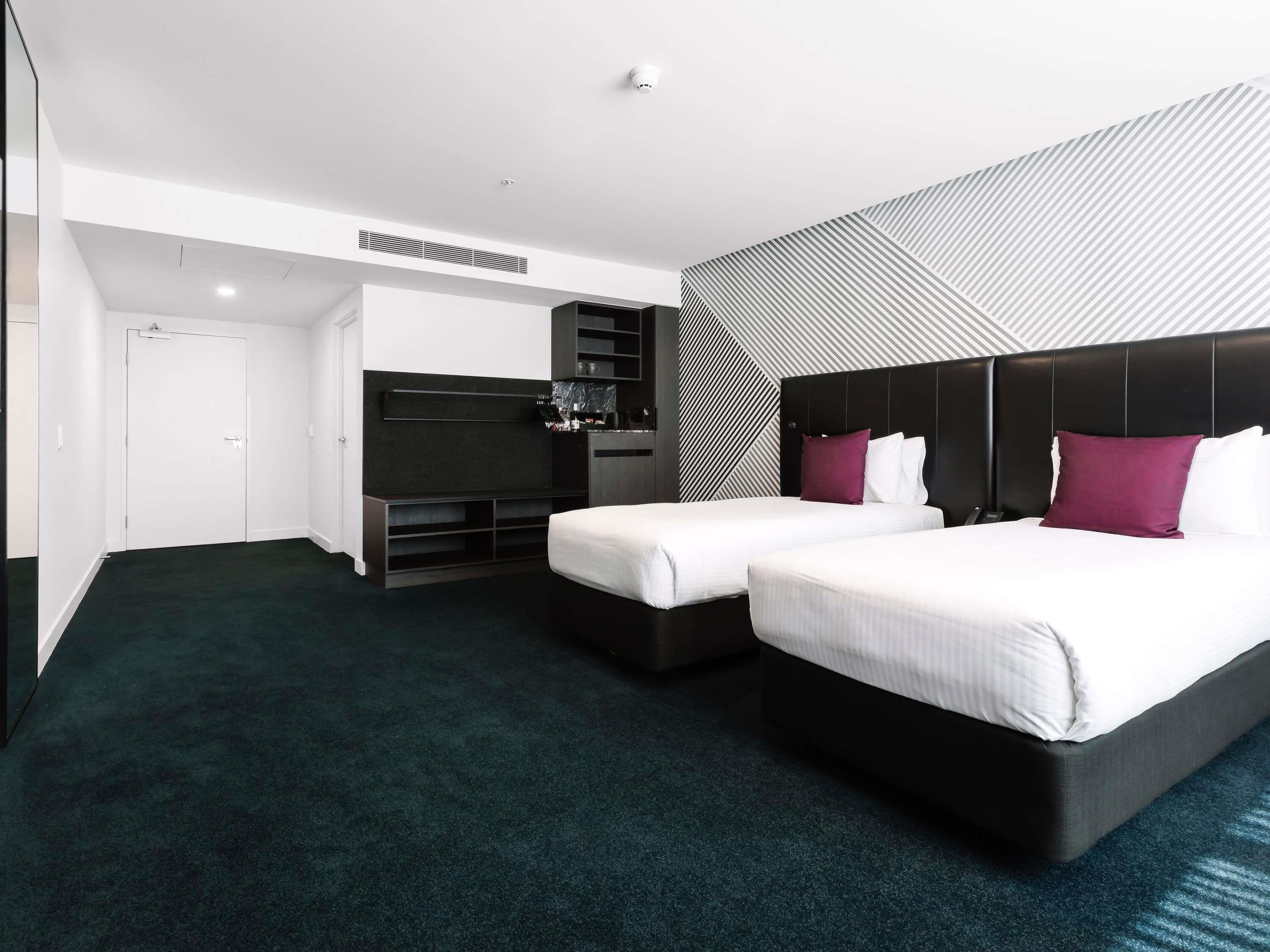 A modern hotel room with two beds, a flat-screen TV, a minibar, and a patterned wall.