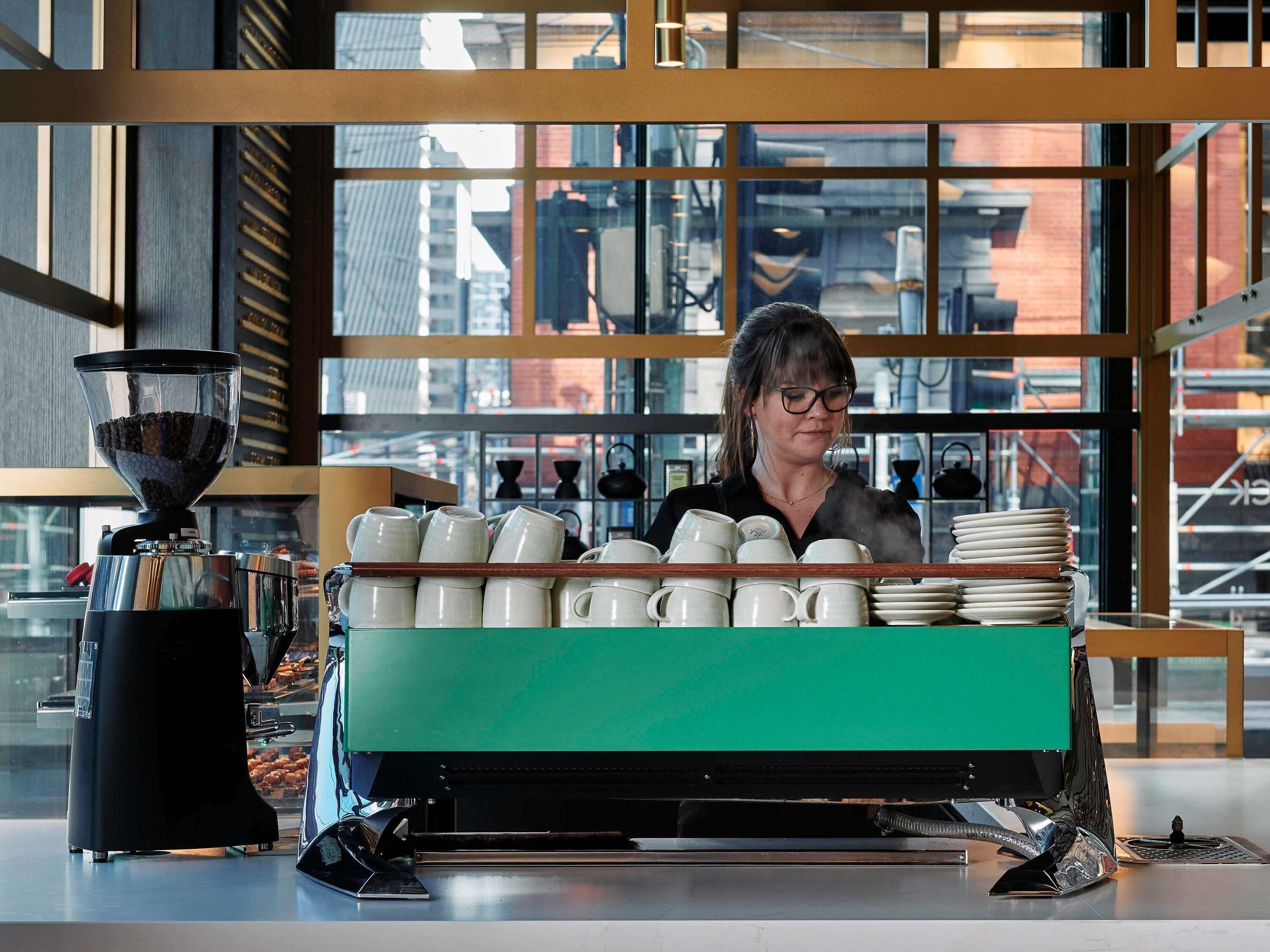Een koffiezaak interieur met een koffieapparaat, een stapel witte mokken, en een uitzicht op de stad.