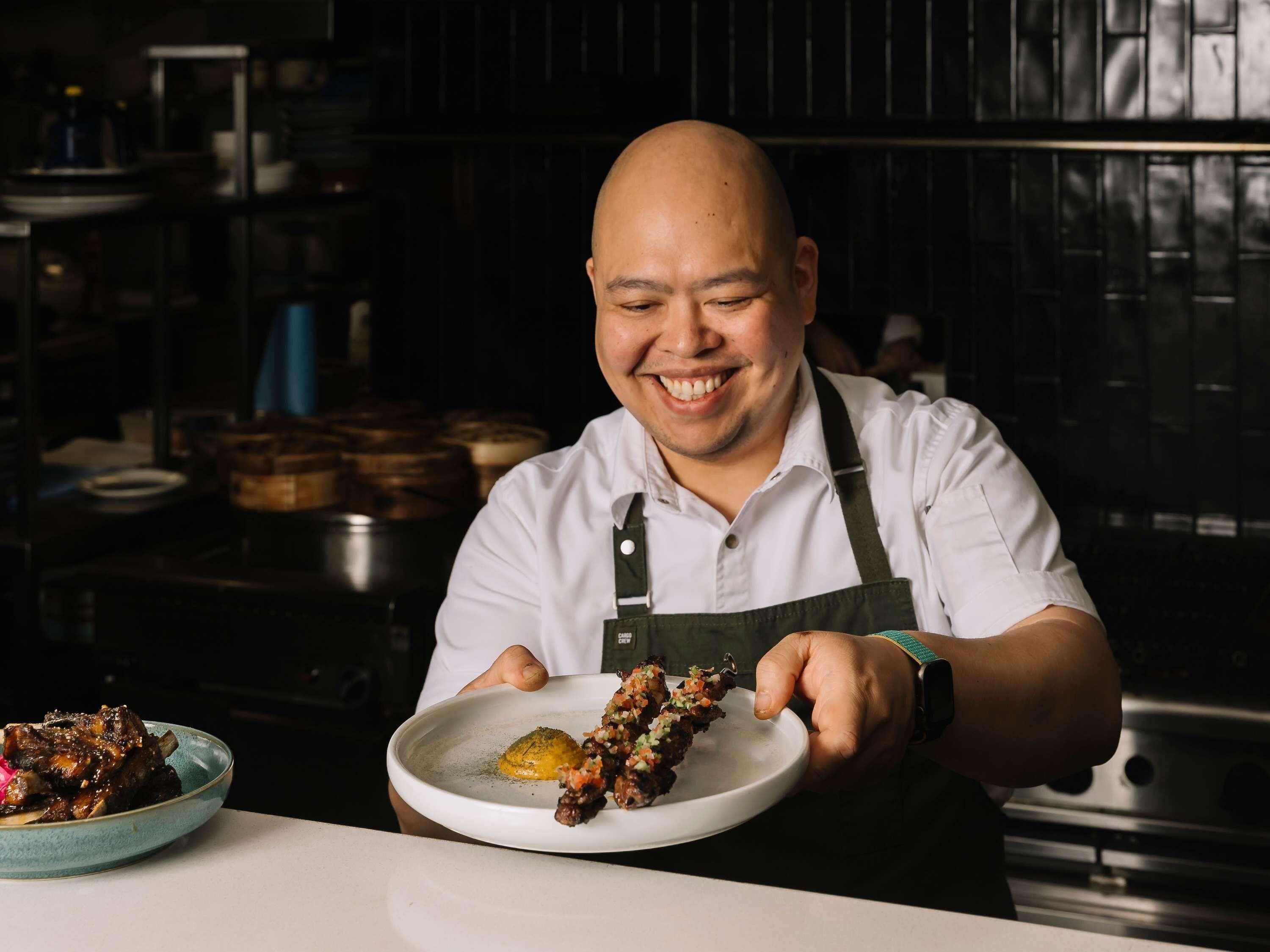 Un chef sosteniendo un plato de comida, en una cocina con estantes y equipo culinario al fondo.