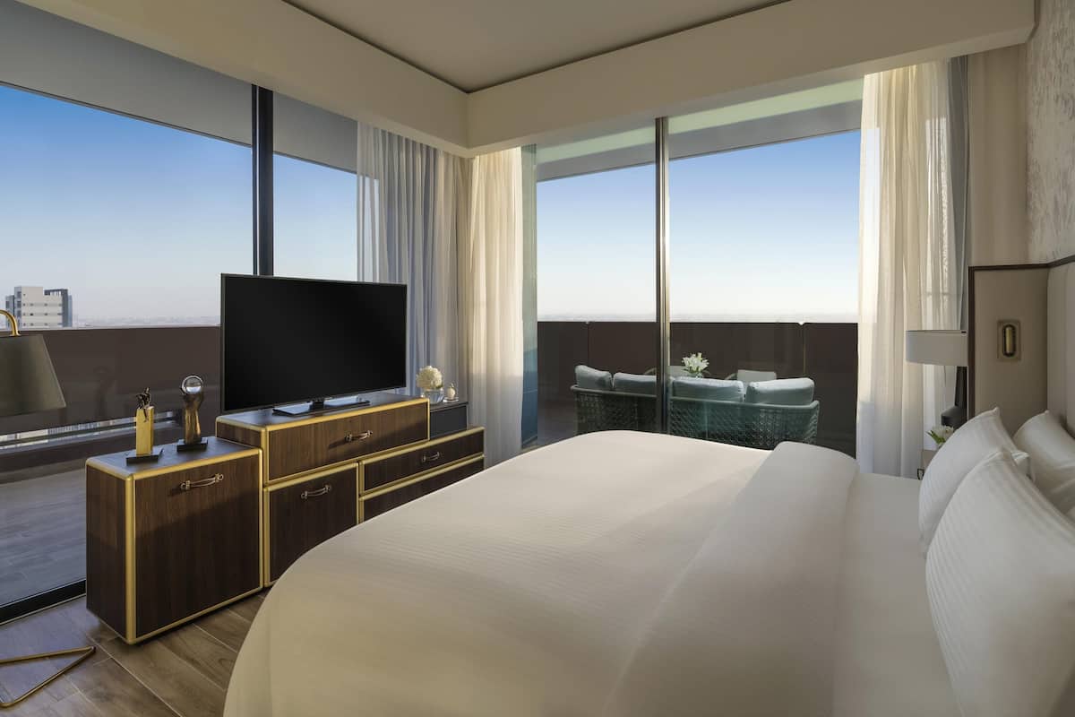 A modern hotel room with a large bed, a flat-screen TV, a wooden dresser, and a balcony with a view of the city.