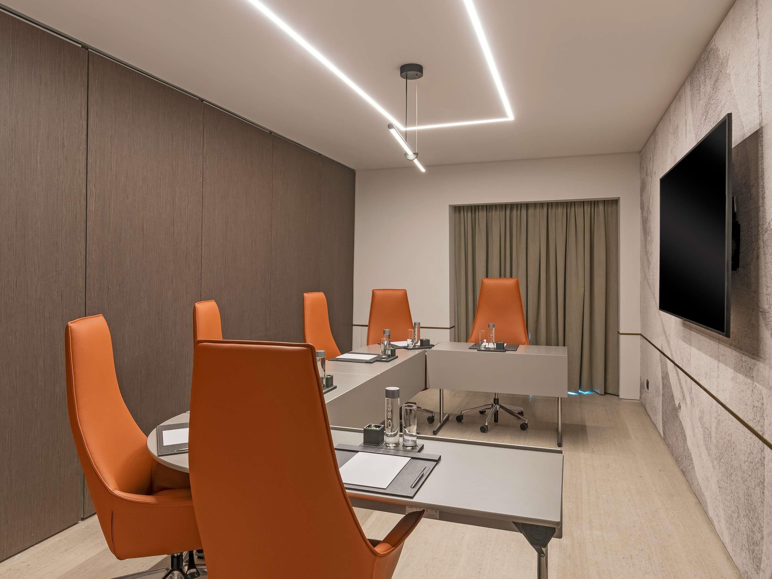 Un salón de conferencias con una larga mesa, cuatro sillas anaranjadas, un televisor de pantalla plana y una lámpara de techo moderna.