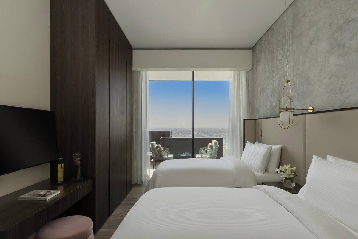 Habitación de hotel con dos camas, televisión, escritorio y balcón con vistas a la ciudad.