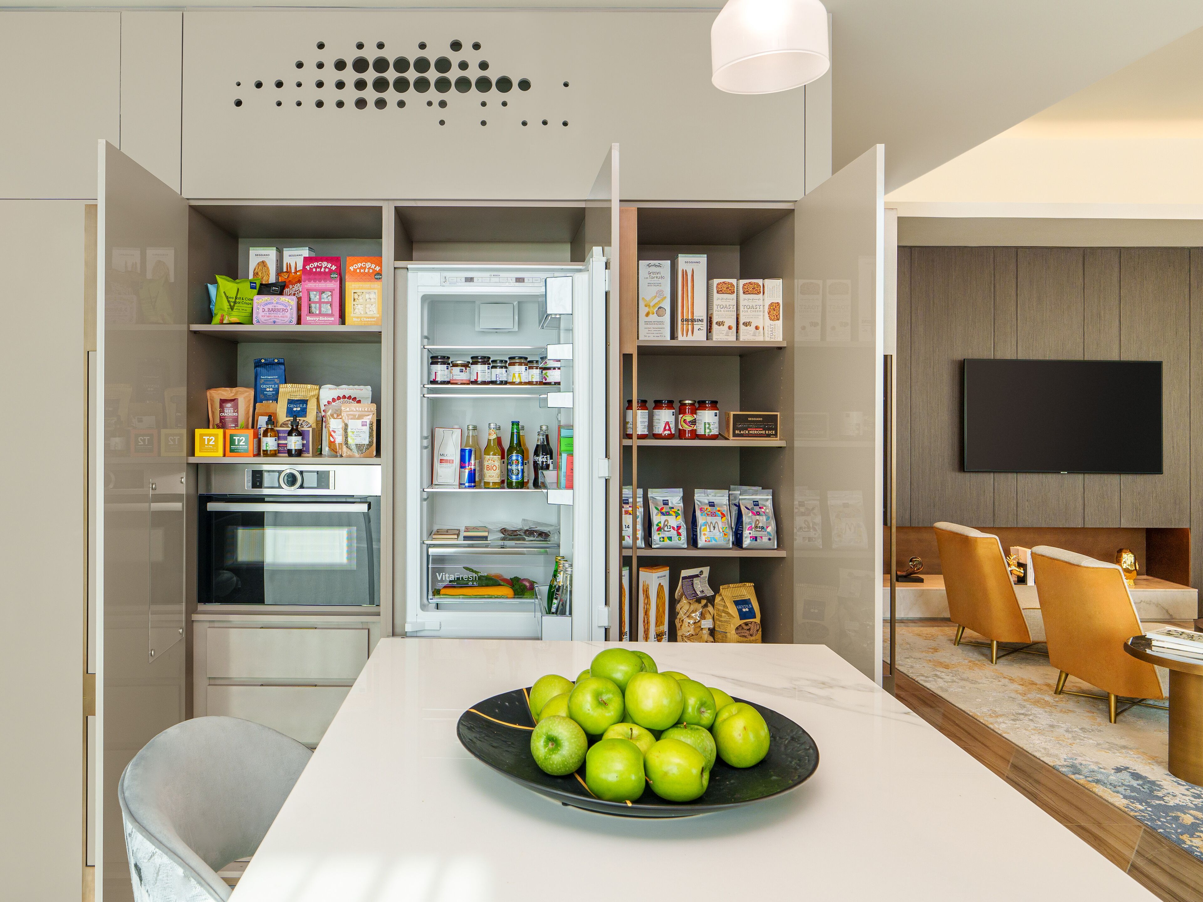 A modern kitchen with a built-in fridge, oven, and microwave. There is a dining table with a bowl of green apples and a TV mounted on the wall.