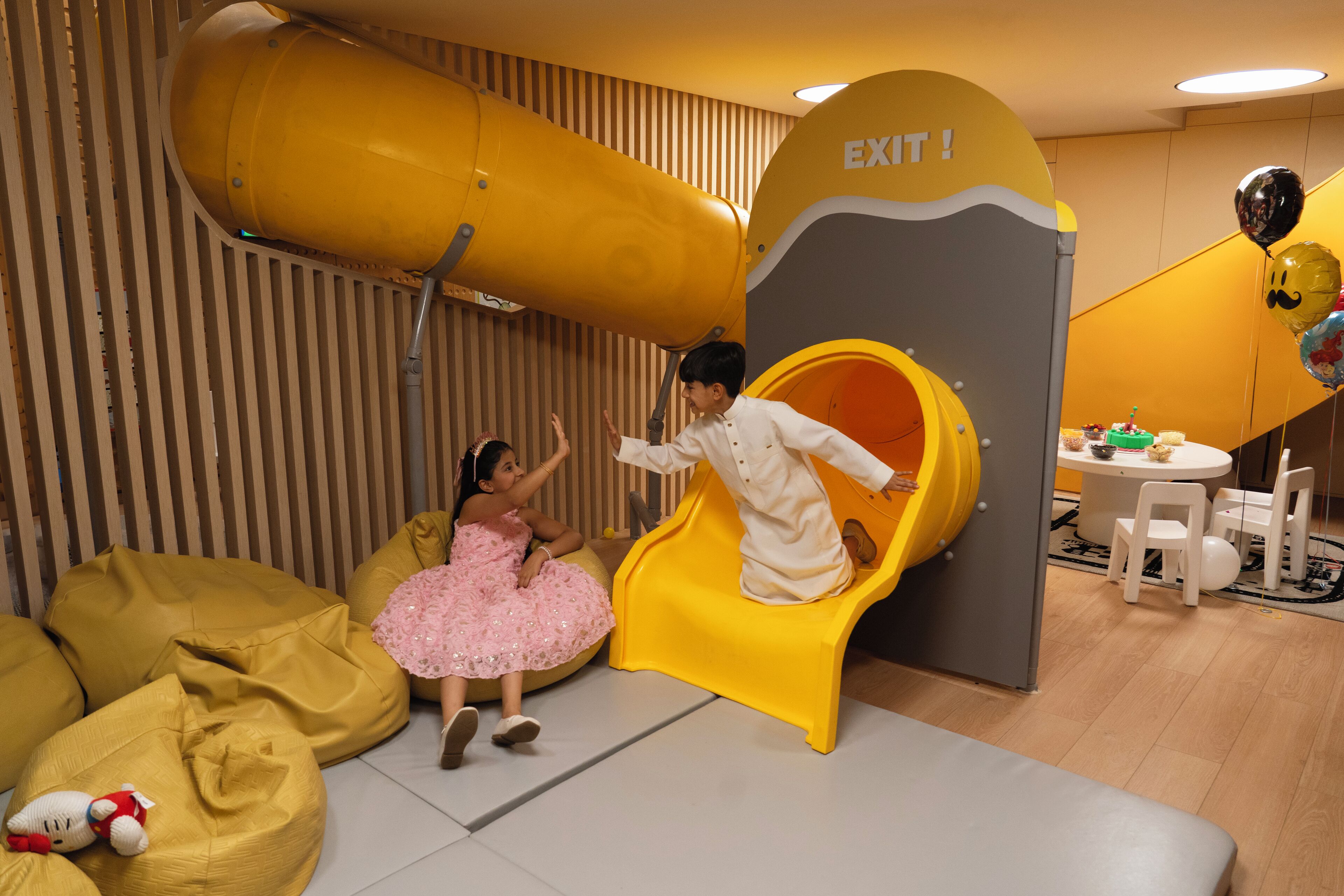 A play area with a yellow slide and a child sliding down. There are bean bags and a table with chairs in the background.
