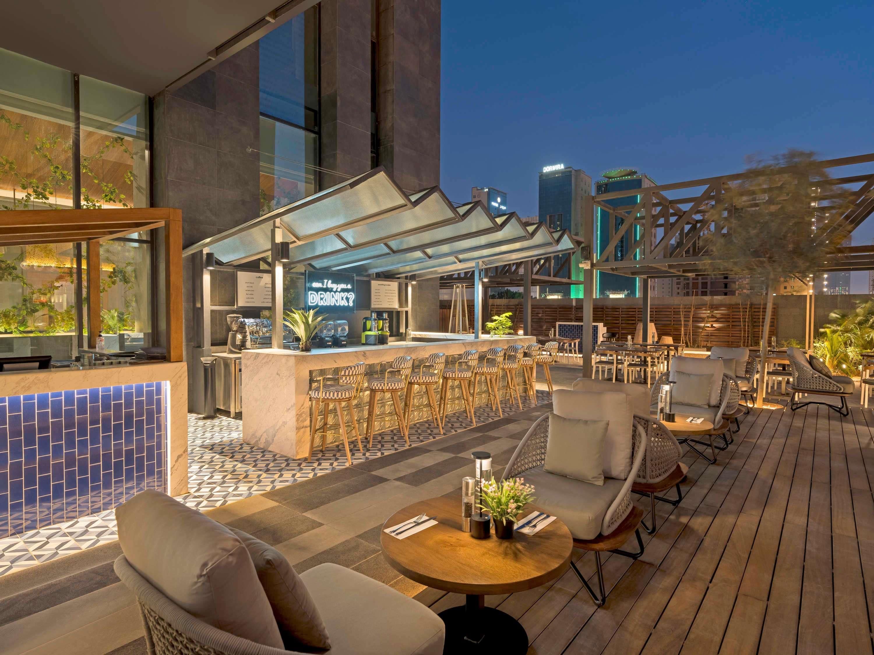 An outdoor bar area with seating, a neon sign, and a cityscape view at dusk.