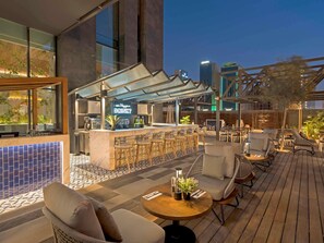An outdoor bar area with seating, a neon sign, and a cityscape view at dusk.
