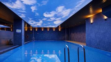 Une piscine intérieure moderne, avec une vue dégagée sur le ciel grâce à une verrière, entourée de murs sombres et dotée d’un éclairage minimaliste.