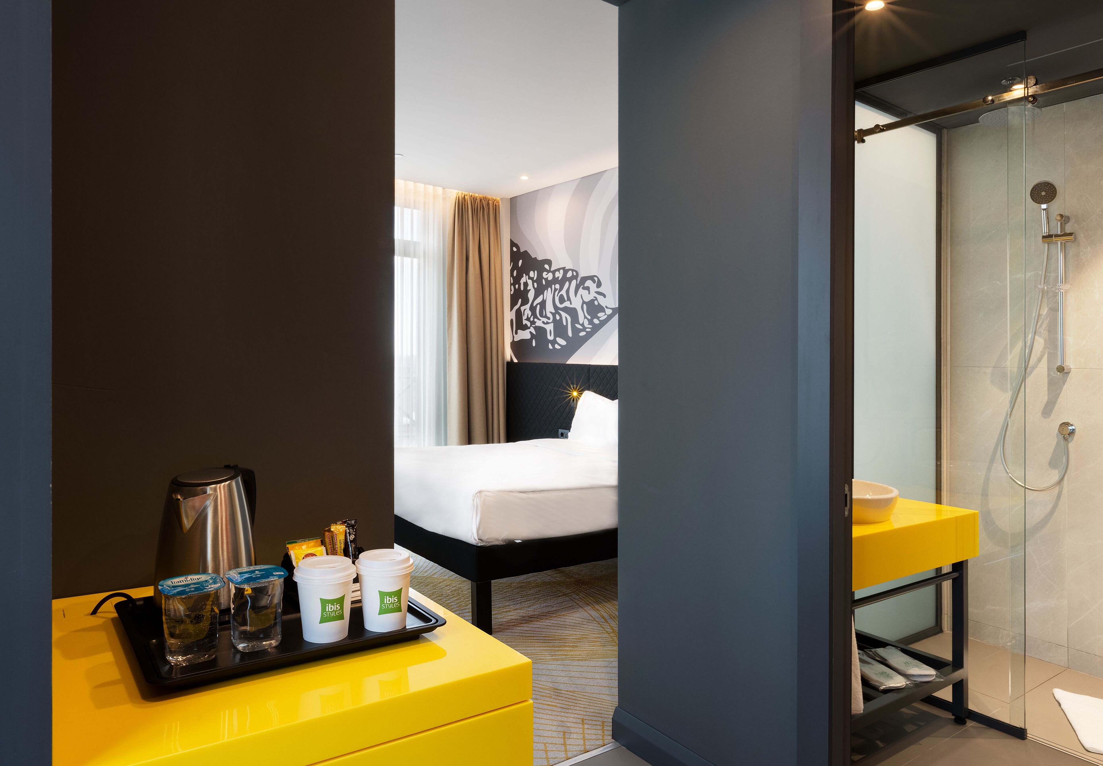 A hotel room with a bed, a bathroom with a shower, a yellow bedside table with a tray of toiletries, and a wall with abstract art.