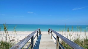 Ein hölzerner Steg führt zu einem Sandstrand mit klarem, blauem Wasser.