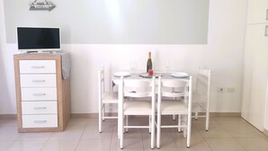 Ein moderner, minimalistischer Raum mit Flachbildfernseher auf einer Holzkommode, einem Esstisch mit weißen Stühlen und einem kleinen, an der Wand befestigten Schild darüber.