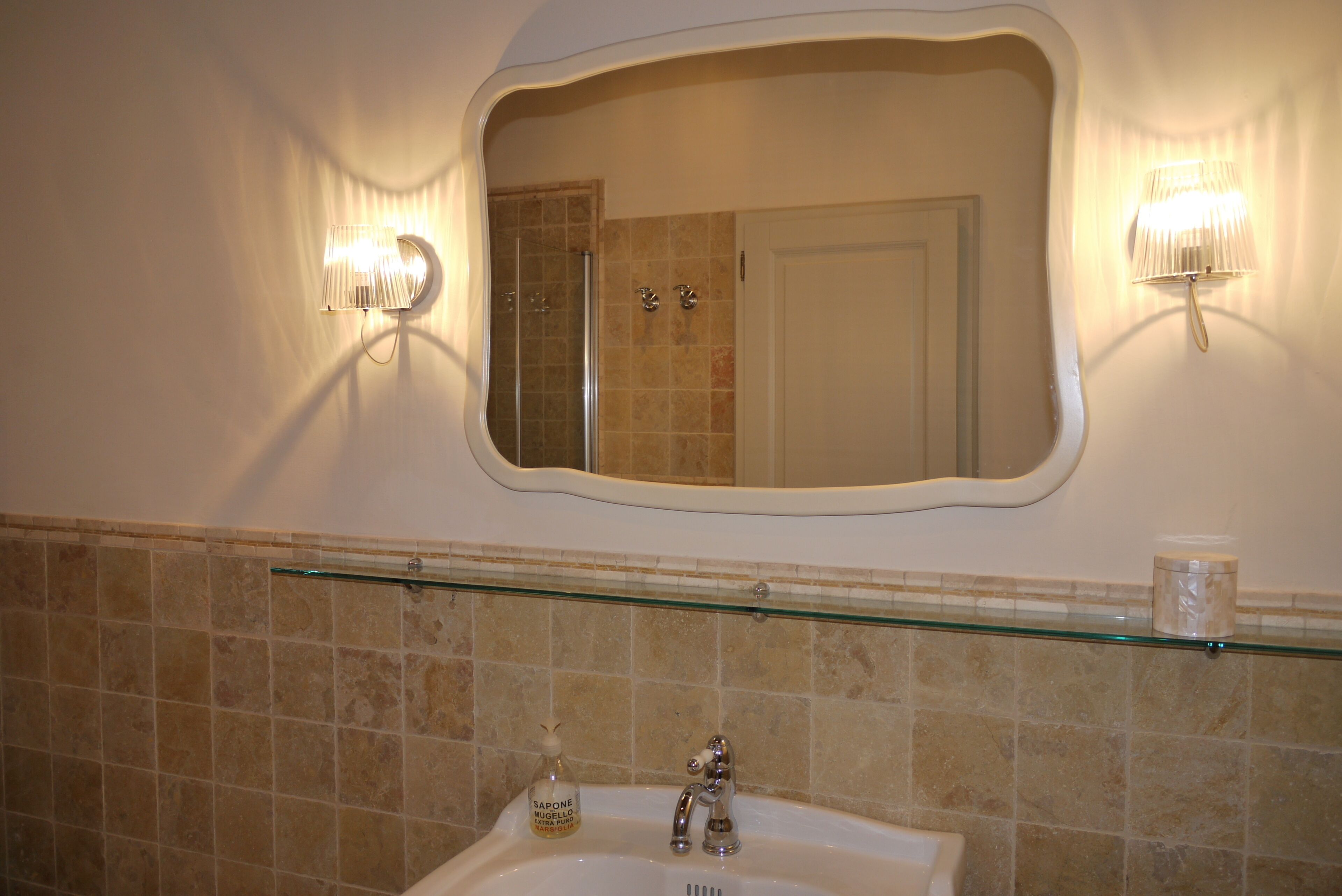 A bathroom with a glass shelf above a sink, a mirror with wall sconces, and a shower area.