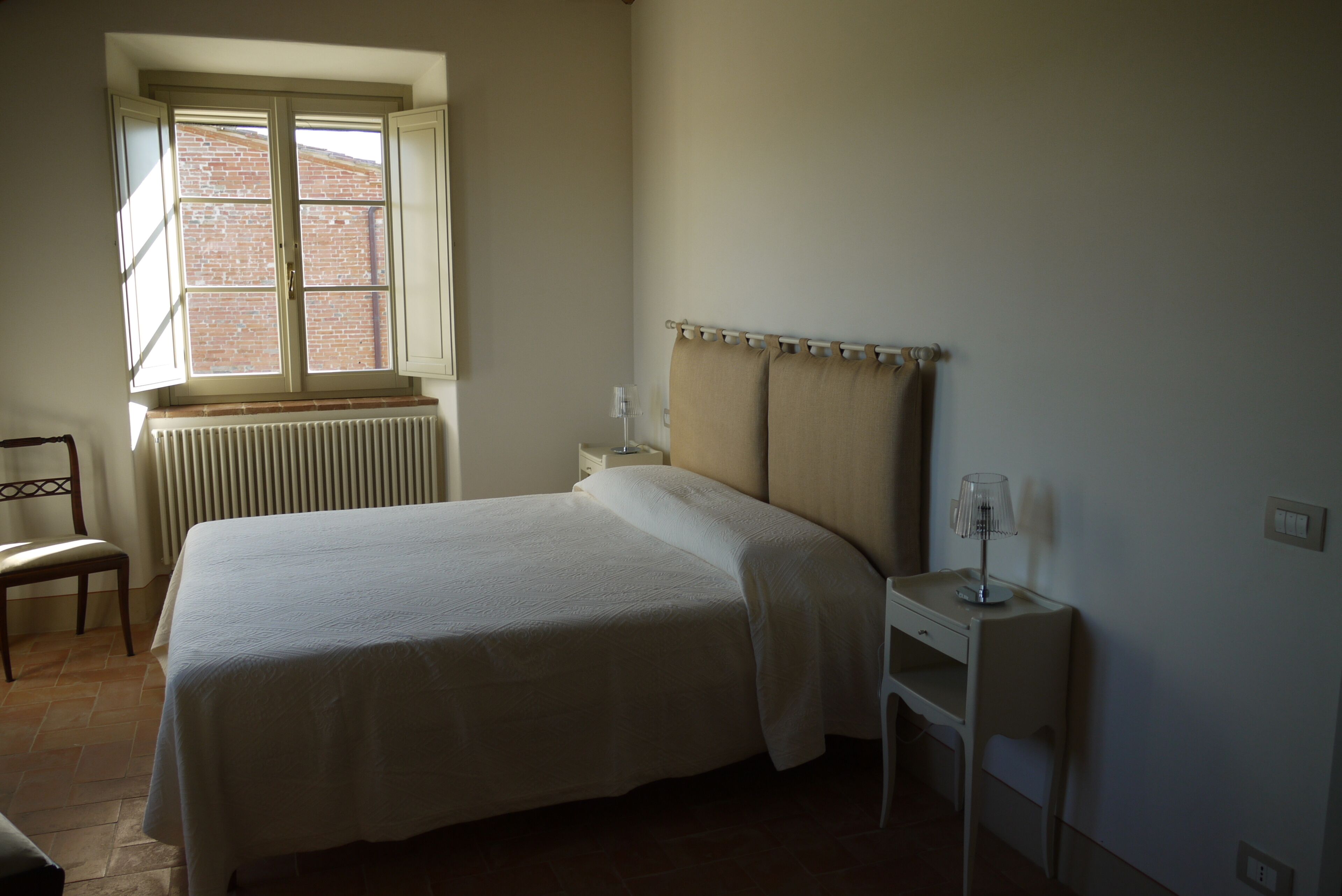 A bedroom with a bed, a nightstand, a chair, a window, and a radiator.
