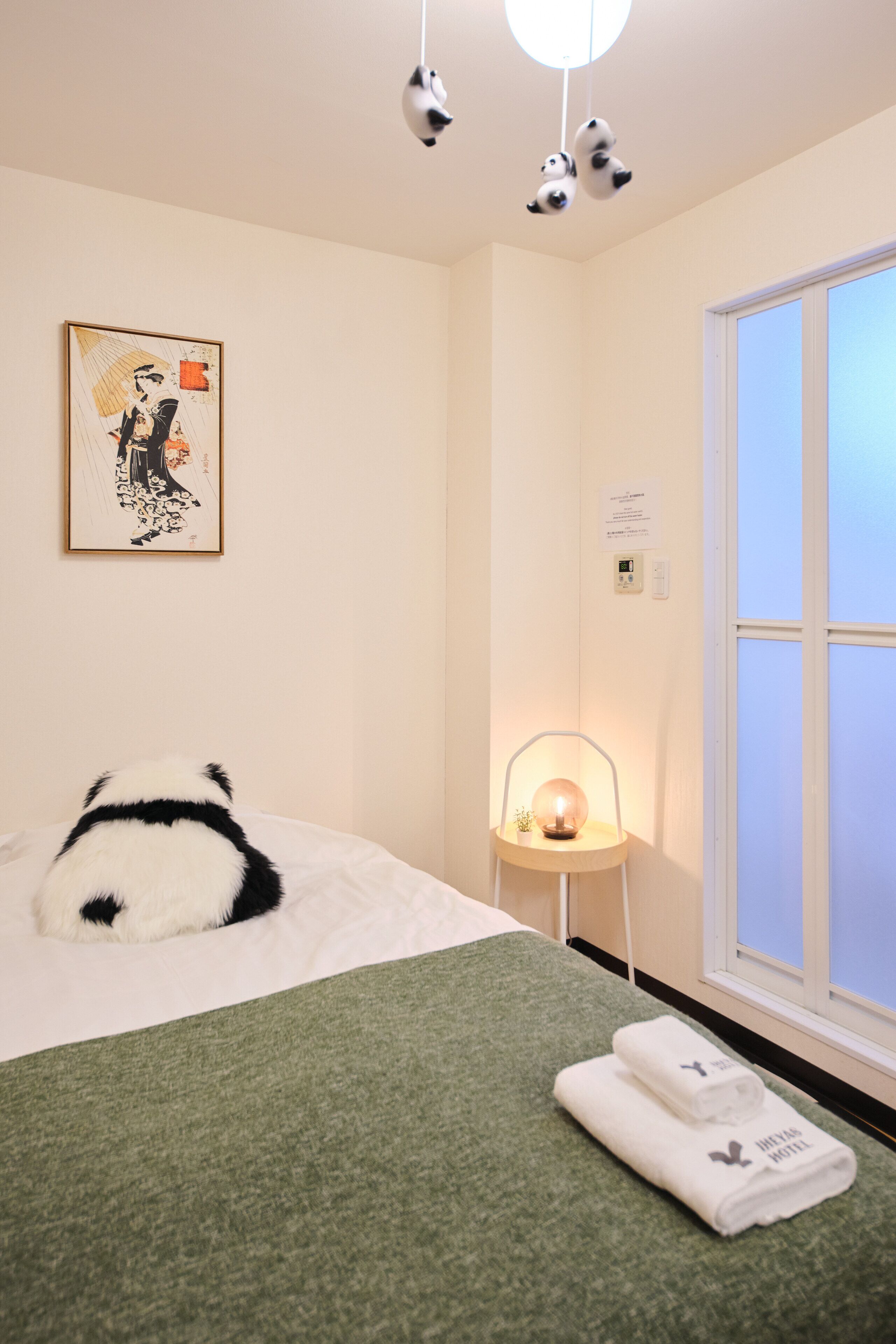 A hotel room with a bed, a bedside table with a lamp, a window, and a panda plush toy on the bed.
