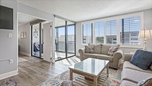A spacious living room with a glass door leading to a balcony, a beige sofa with patterned cushions, a glass coffee table, and a view of the city through large windows with blinds. - 🌺Remodeled Coastal Retreat With Beach Gear 🐶 Loggerhead Cay 463🐠 (Sanibel)