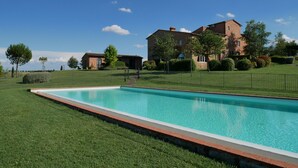 Una piscina rectangular con borde de césped, rodeada por una valla, y un edificio de ladrillo grande con varias ventanas y una torre al fondo.