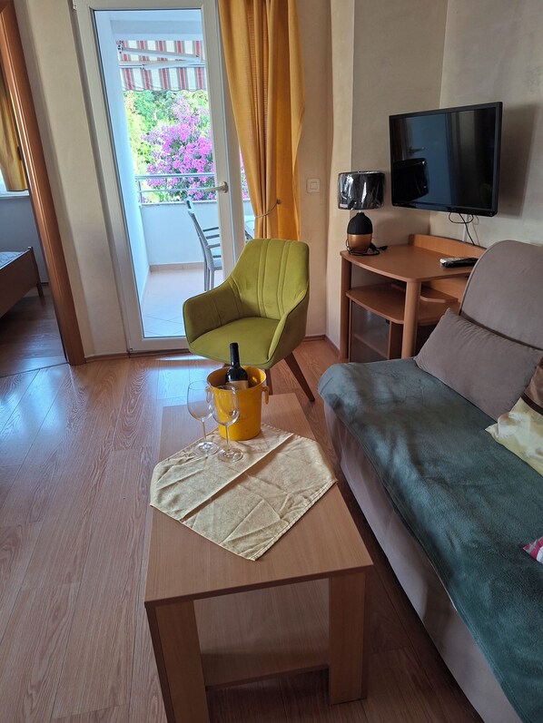 Uma sala de estar com uma poltrona verde, uma mesa de centro de madeira, um sofá, uma TV e uma varanda com vista para flores.