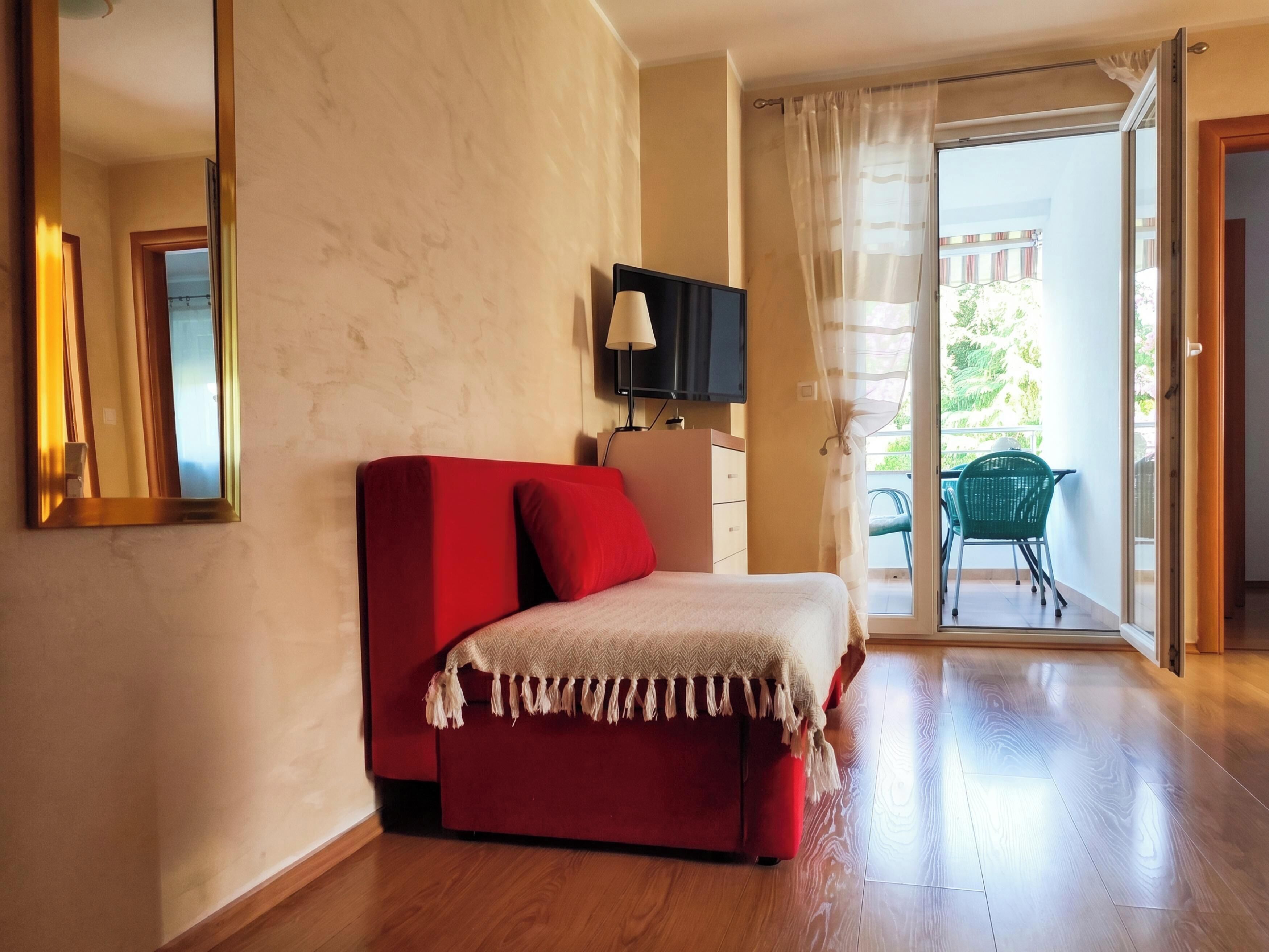 A room with a red sofa, a television on the wall, a sliding glass door leading to an outdoor area with a chair.