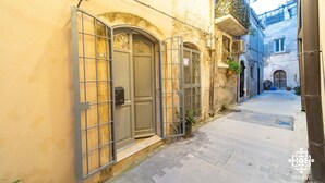 A narrow street with a building featuring a metal gate, a door with a decorative arch, and a small balcony with plants. - Casa Sophia (Syracuse)