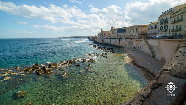 A coastal view with clear waters, a rocky shoreline, and historic buildings along the promenade. - Casa Sophia (Syracuse)