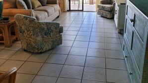Ein großzügiger Wohnbereich mit einer Couch, Sesseln und einem Esstisch. Eine große Schiebetür öffnet sich zu einem Balkon mit Blick aufs Meer.