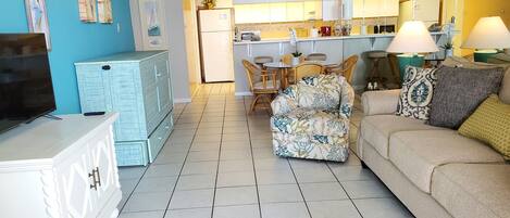 Una sala de estar con televisor, sofá, sillón floral y una cocina al fondo.
