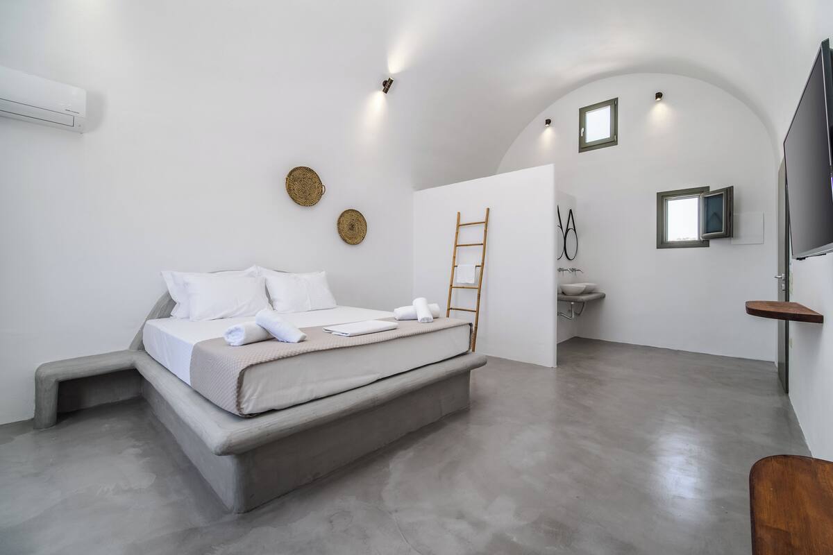 A modern bedroom with a bed, a small table, a TV, and a bathroom with a sink and mirror.