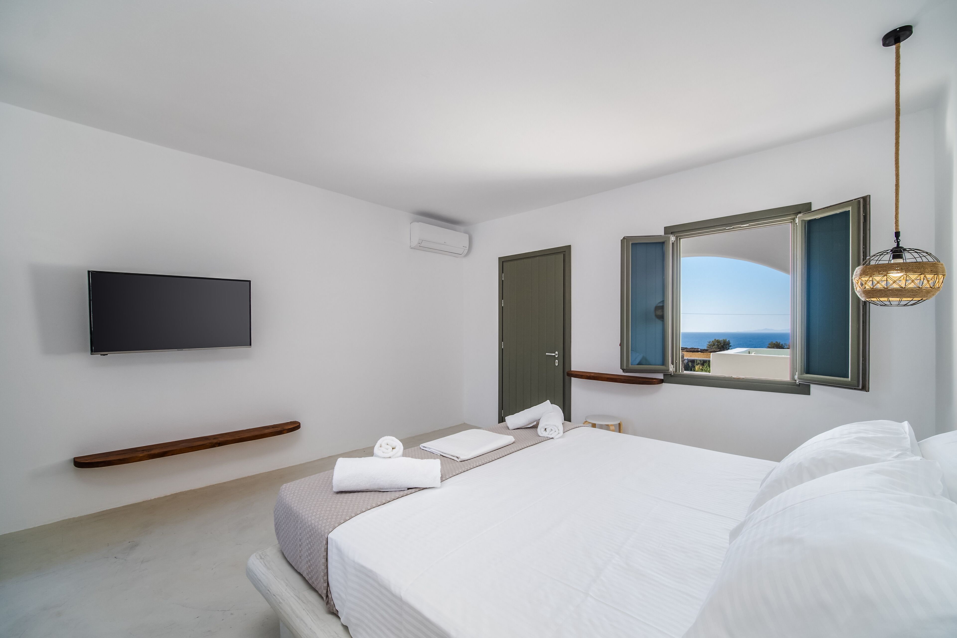 Une chambre d’hôtel moderne dotée d’un grand lit, d’un téléviseur à écran plat fixé au mur, d’une étagère en bois et d’une fenêtre donnant sur la mer.