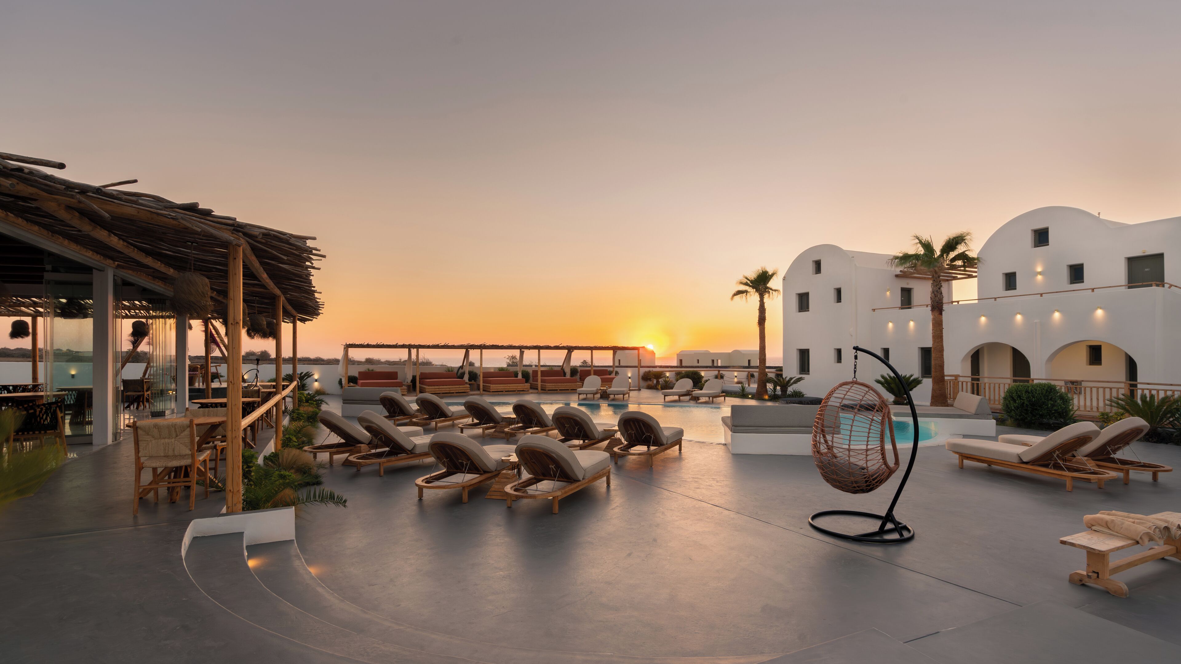 Área de piscina en la azotea con sillones de descanso y una silla colgante, con vista a una puesta de sol.