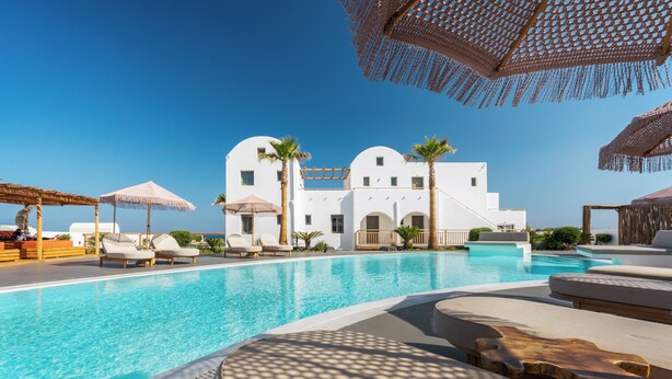 Un área de piscina con sillones, sombrillas y un edificio blanco con arcos.