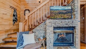 A cozy cabin interior with a stone fireplace, wooden staircase, and a television mounted above it. - 2Kings!Jacuzzi*Fireplace*Pool*Pets ok (Sevierville)