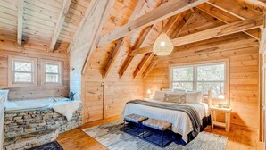 A cozy wooden cabin bedroom with a stone fireplace, a bed with a blue and white bedspread, and a hanging light fixture. - 2Kings!Jacuzzi*Fireplace*Pool*Pets ok (Sevierville)