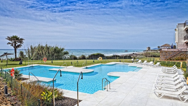 A pool area with a view of the ocean, surrounded by lounge chairs and a building with a stone facade. - Casares Del Mar (Casares)