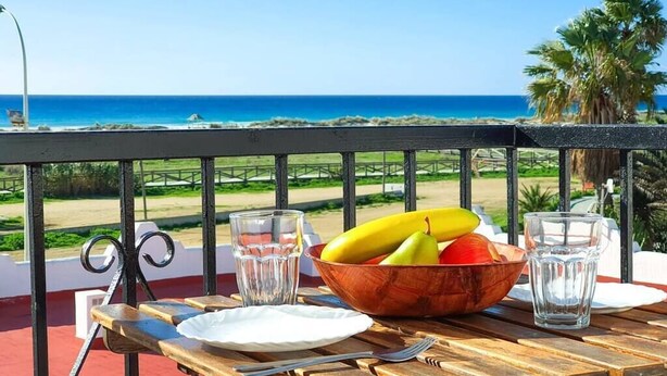 Ein Holztisch mit Obstschale, zwei Gläsern und Besteck auf einem Balkon mit Blick auf einen Strand.