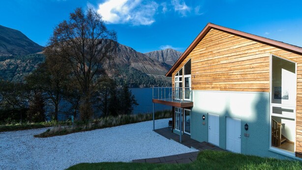 A modern wooden cabin with a balcony overlooking a lake and mountains.