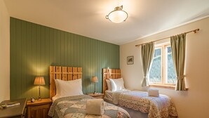 A bedroom with two beds, wooden headboards, a nightstand with a lamp, a window with curtains, and a wooden floor.