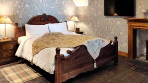 Een traditionele slaapkamer met een houten bed, twee nachtkastjes met lampen, een open haard en bloemenbehang.