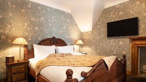 A traditional bedroom with a wooden bed, two bedside tables with lamps, floral wallpaper, and a flat-screen TV mounted on the wall. - Rhododendron Suite -With views of the rhododendron trees on the hillside (Inverness)