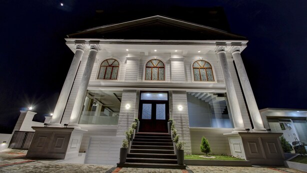 Un edificio grande y blanco de dos pisos con fachada simétrica, ventanas amplias con marcos decorativos y una entrada central con escaleras.