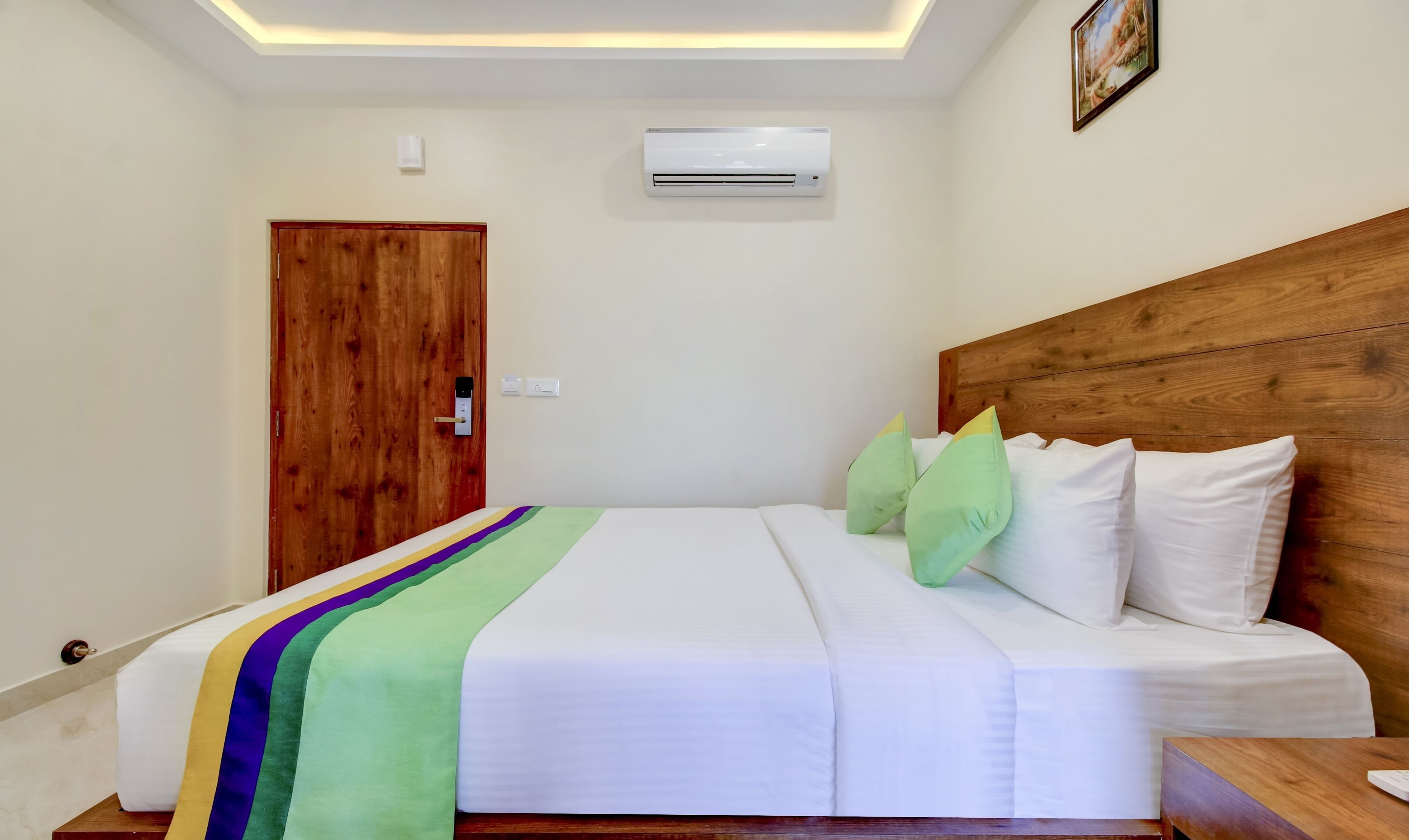 A hotel room with a wooden headboard, a bed with white bedding and green pillows, an air conditioning unit on the wall, and a wooden door.