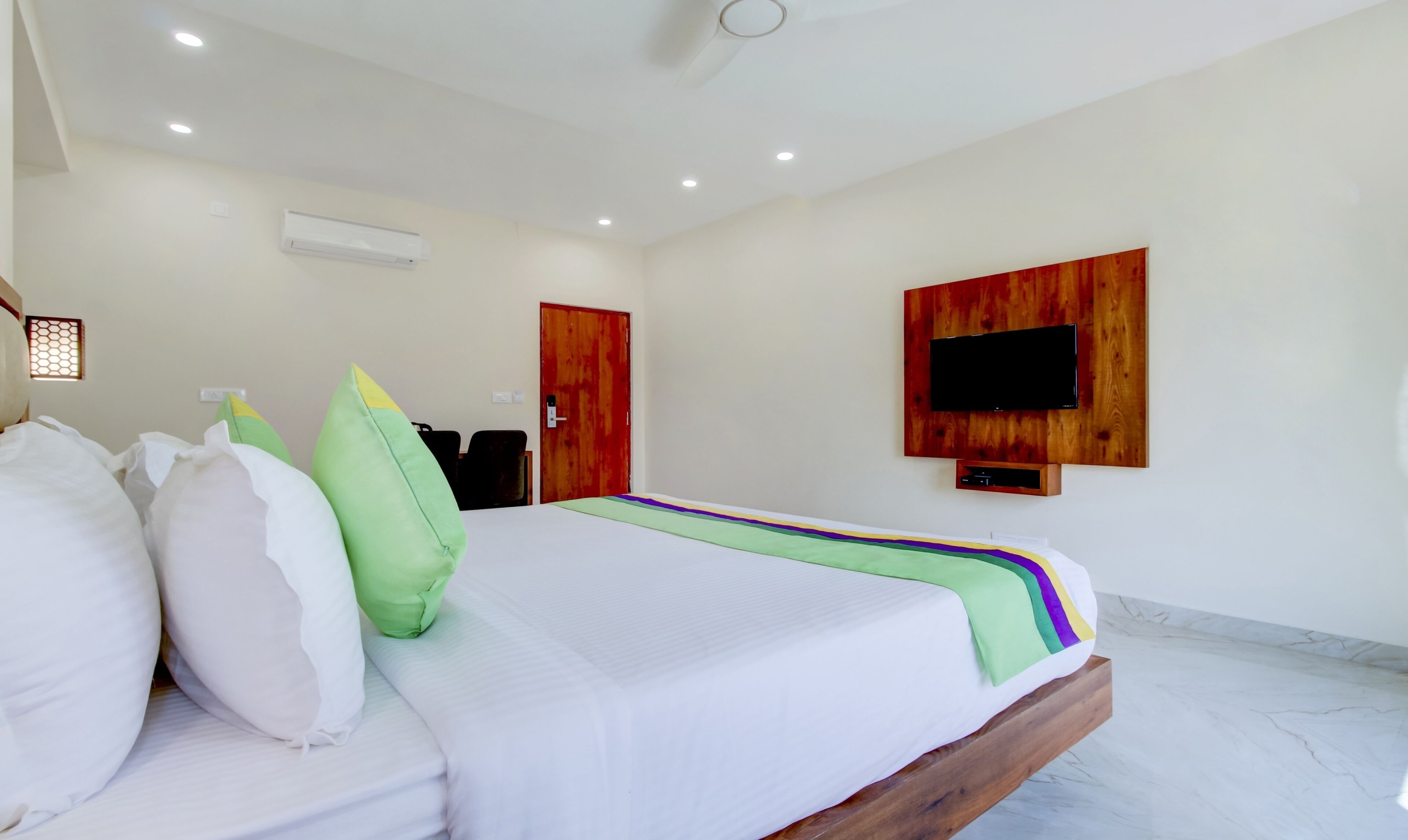 Een hotelkamer met een bed, kussens, een aan een houten paneel bevestigde televisie en een plafondventilator.