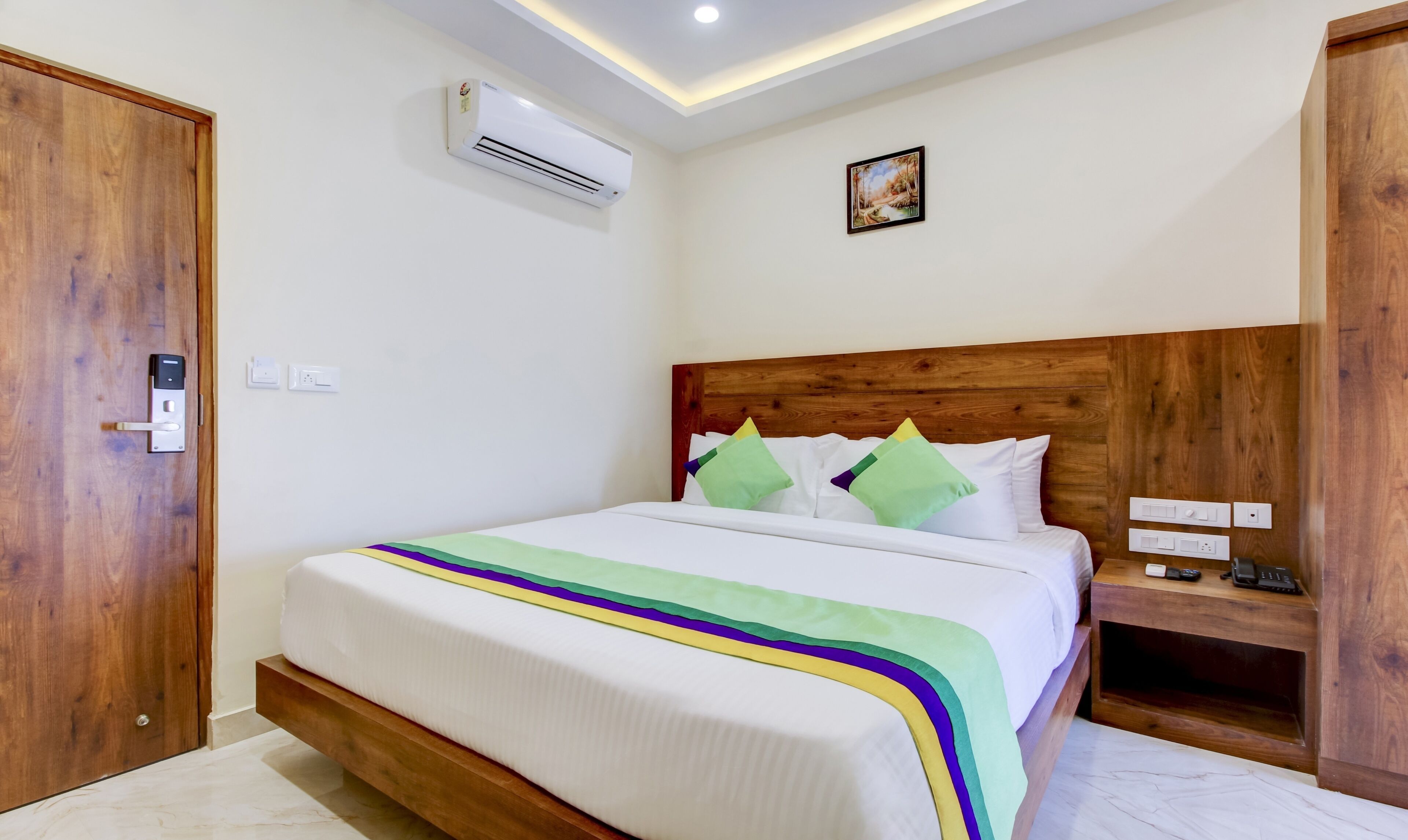 A neatly arranged bedroom with a wooden headboard, a bed with white bedding and colorful pillows, an air conditioning unit on the wall, and a small bedside table with a phone and charger.