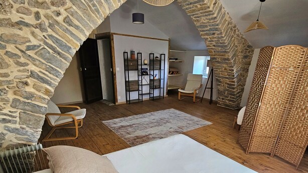 A cozy attic bedroom with a stone wall, a bed with pillows, a chair, a small table, and a hanging light fixture.