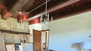 A living room with a sofa, armchairs, a coffee table, a chandelier, a lamp, and a kitchen area with a stone wall and wooden ceiling.