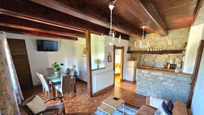 A spacious living area with a stone wall, wooden ceiling beams, a dining area with a table and chairs, and a kitchen with a stone countertop.